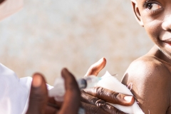 Excited little African boy with hopeful eyes, raised eyebrows and an uneasy smile on his face looking at his pediatrician who is going to inject an intramuscular solution into his arm